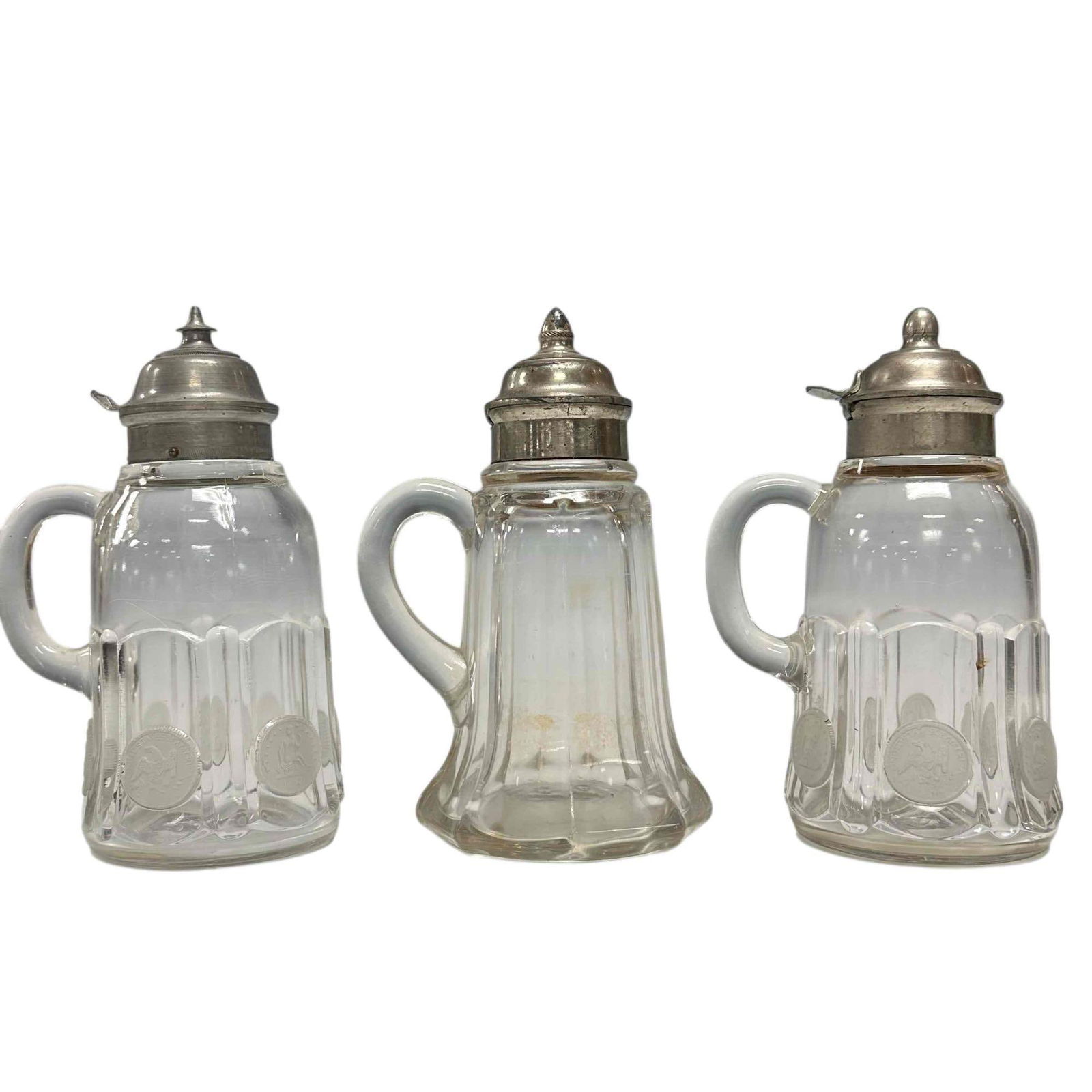 Early American Pattern Glass Syrup Pitchers (3): Late 19th early 20th century Early American Pattern Glass syrup pitchers with pewter lids. Two of the three having "Silver Age" U.S. coin pattern on glass panels. Measuring approx. 7" tall. In age app