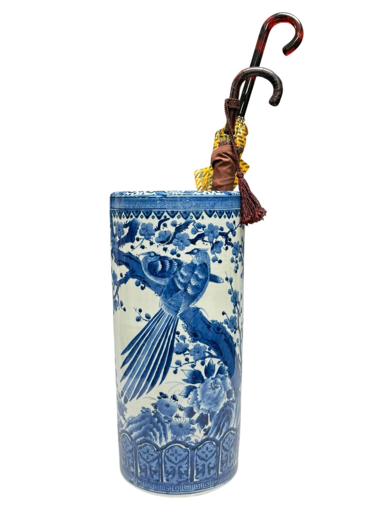 Japanese Blue and White Porcelain Umbrella Stand with Umbrellas: Porcelain blue and white umbrella stand depicting birds perched upon tree branch surrounded with blossoming foliage measuring 18" tall by 8.5" diameter. Containing two vintage umbrellas with one havin