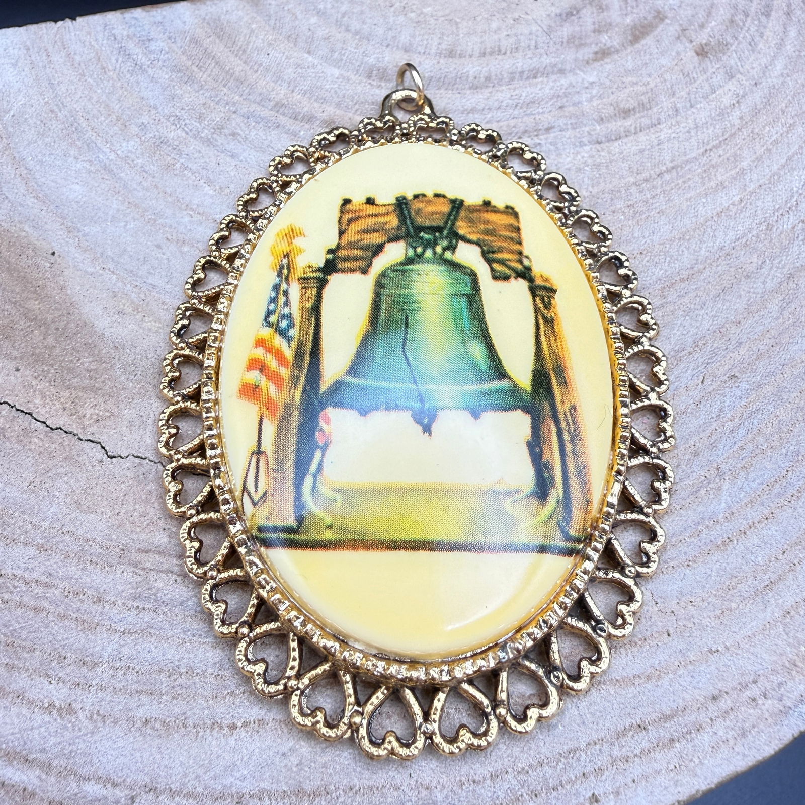 Vintage Liberty Bell Cameo Pendant, Gold Tone: Oval cameo-style pendant featuring a patriotic image of the Liberty Bell with American flag, set under a domed clear cabochon (appears resin or plastic) in an ornate gold-tone metal frame with heart-m