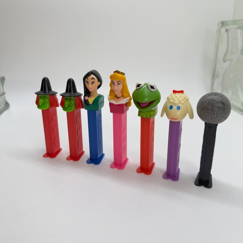 Group of 7 Character PEZ Dispensers: Group of seven character PEZ candy dispensers featuring mixed pop-culture and novelty themes, including two witch figures, Pocahontas, Princess Aurora, Kermit the Frog, a lamb, and a black dispenser t