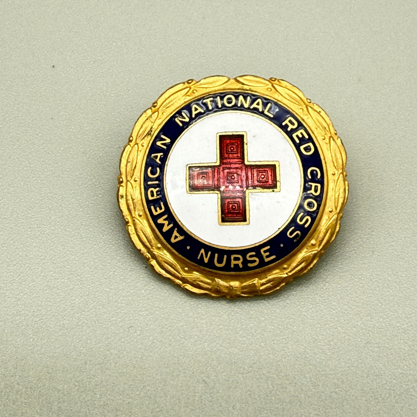 Vintage American Red Cross Nurse Enamel Pin Badge: Vintage gold-tone and enamel nurse badge from the American National Red Cross. Round laurel-wreath form with deep blue enamel border reading “American National Red Cross Nurse” surrounding a r