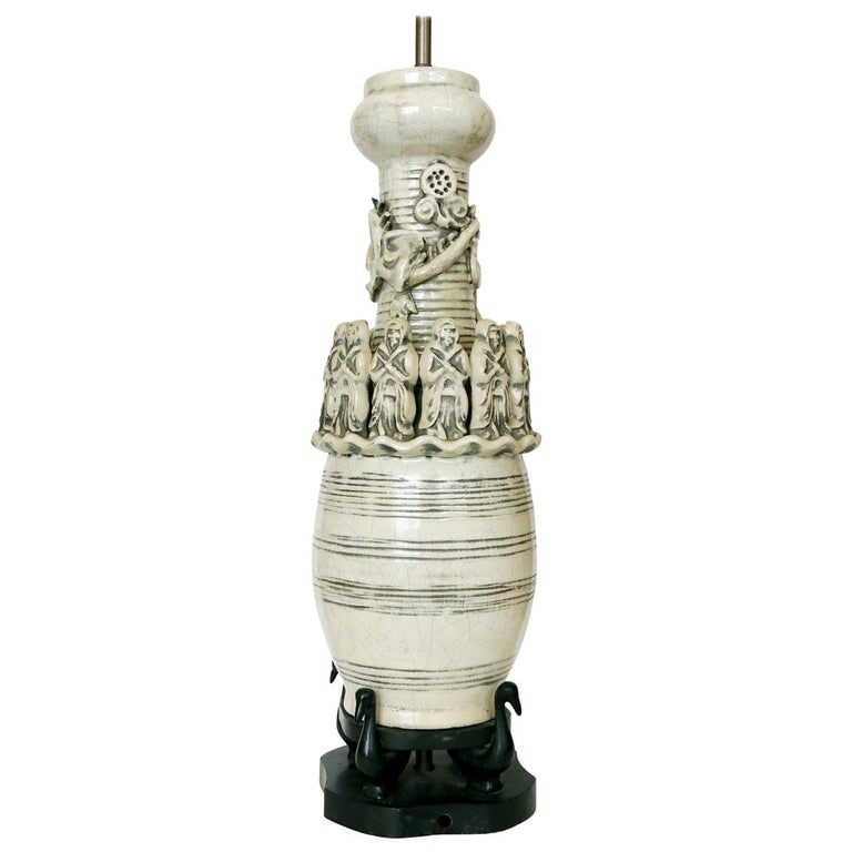 Art Pottery Ceremonial Monks with Dragon Table Lamp: The work is a white art pottery lamp of a cylindrical shape with a wood base. The lamp is decorated with 12 monks that surround a dragon. The lamp also has decorative, horizontal patterns and a crackl