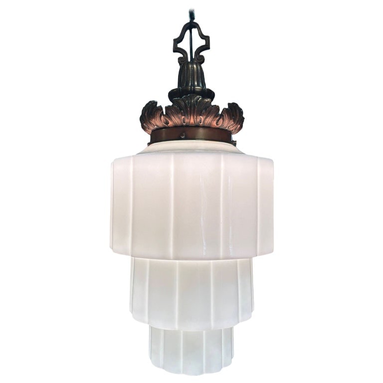 Art Deco Era Ornate Brass Pendant Light w/ Stepped Milk Glass Skyscraper Shade: This 1930s Art Deco pendant light features a tiered Art Deco milk glass shade featuring stepped geometric layers and is paired with an ornate cast brass fitter in a warm bronze finish. The crown showc