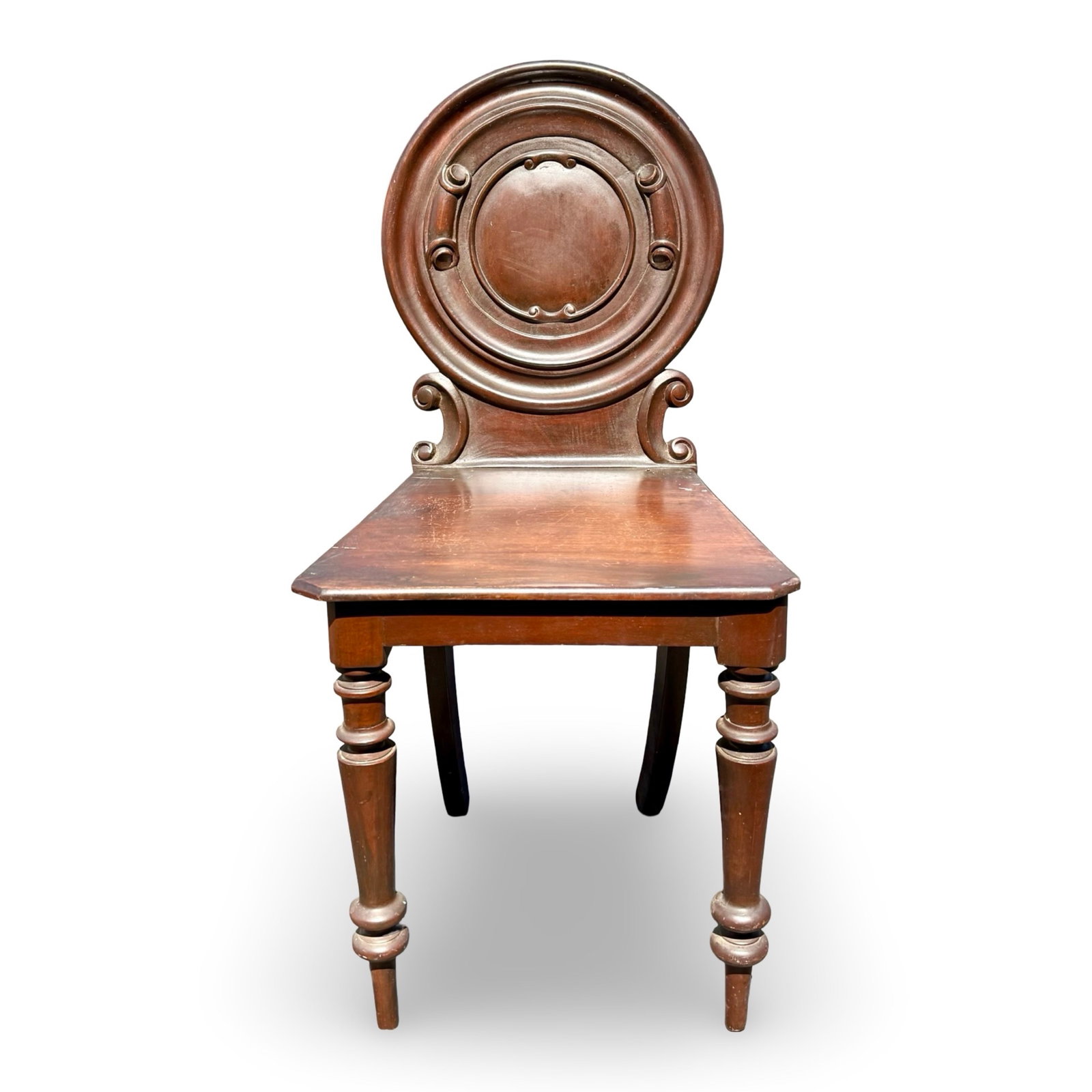 19th Century English Mahogany Target Chair: 19th-century English mahogany hall chair featuring a molded circular target back with carved scroll supports, solid panel seat, turned front legs, and splayed rear legs. The chair displays a warm aged