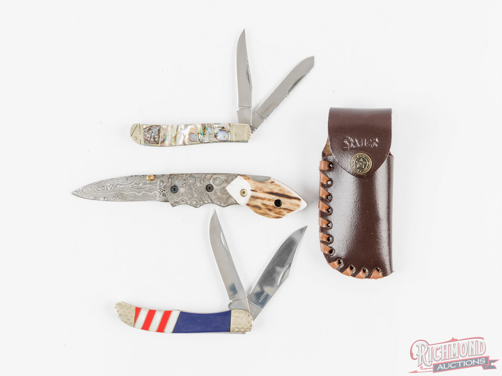 Three Stauer Folding Knives Abalone, Damascus And Stag, USA Stars & Stripes: Trapper two blade with abalone inlay handle, brass liners and nickel bolsters. Measures 3-1/2" closed. Stars & Stripes two blade folder features red, white and blue handles with brass liners and
