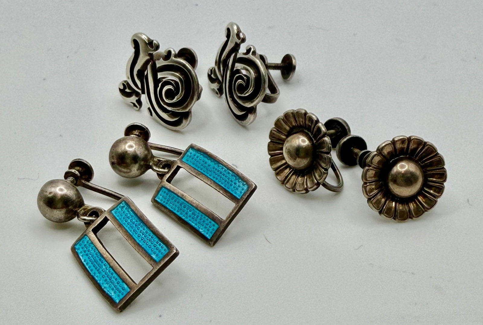 Three Pairs of Vintage Sterling Silver Earrings: Three unique pairs of vintage screw-on sterling silver earrings. The first pair are a dangle style and feature turquoise enameled rectangles. The second pair are an attractive scroll design within a s