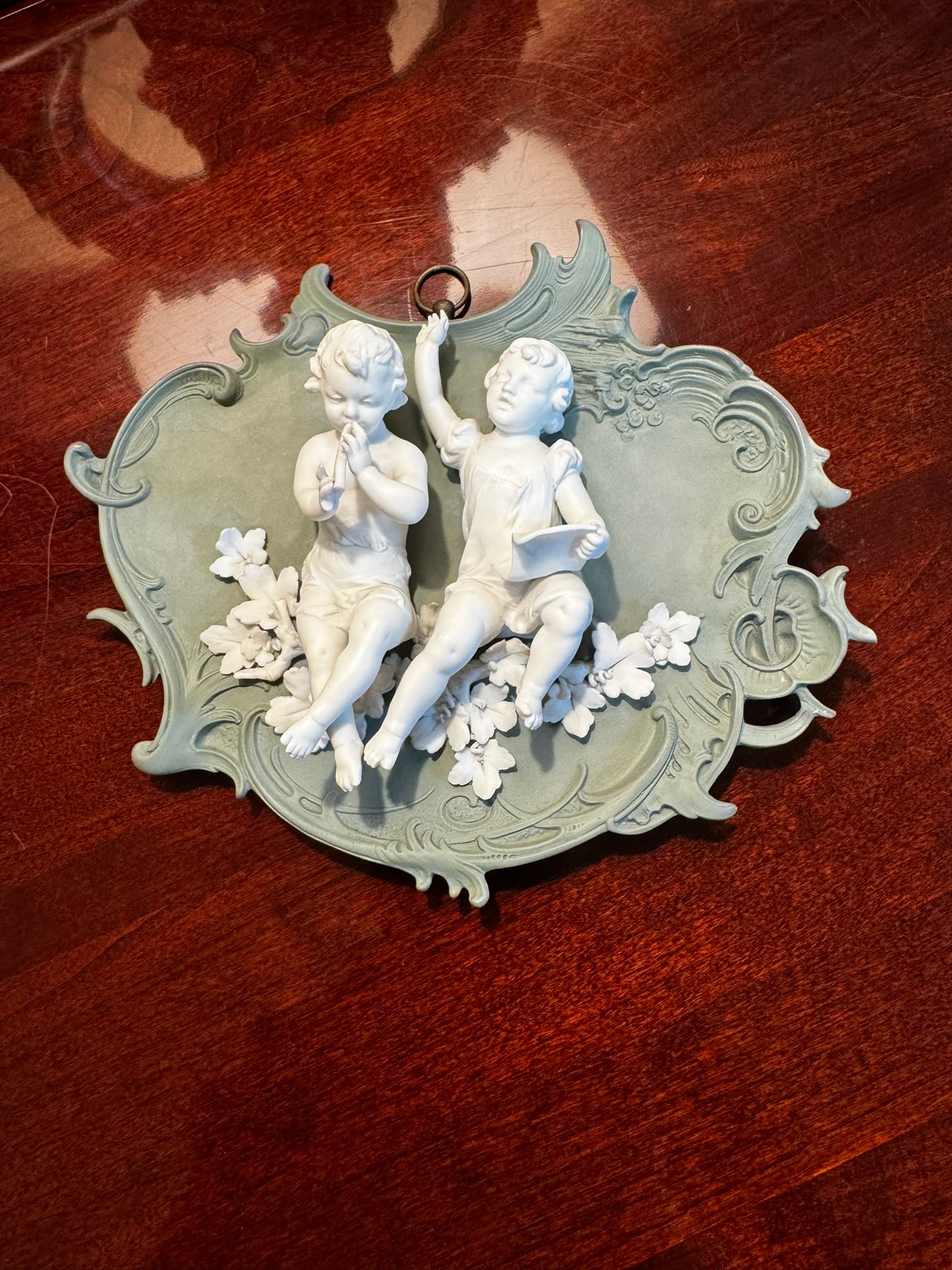 Volkstedt Green Jasperware Cherub Plaque: An ornate vintage wall plaque by Volkstedt featuring high-relief white bisque porcelain cherubs seated among flowers against a classic green jasperware ground.