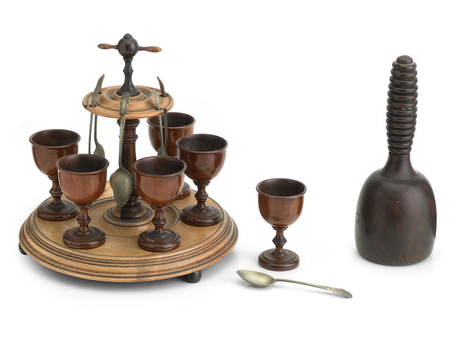 A treen egg stand 19th century: A treen egg stand 19th century Turned in sycamore, mahogany and lignum vitae and formed as a circular moulded stand, six egg cups and a central column, 21cm high, together with a rosewood mallet with