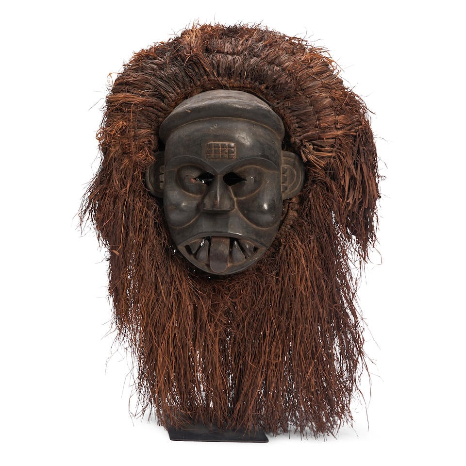 CARVED IBIBIO MASK: CARVED IBIBIO MASK The face with geometric decorations on the forehead and ears, large teeth, and a protruding tongue, surrounded by a mane and beard of grass, on a stand, 27' height, 18' width, 7' de