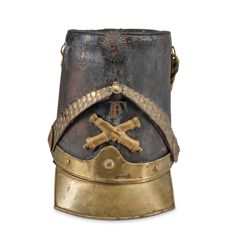 AN AMERICAN MILITIA ARTILLERY SHAKO.: AN AMERICAN MILITIA ARTILLERY SHAKO. c. 1840. Black leather body centering embossed brass crossed cannon below 'F' and a brass scale chin strap and pressed leather cockade, all above a brass visor, br