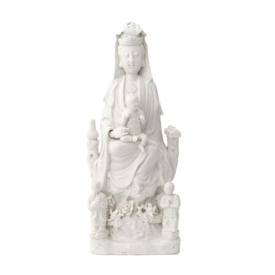 A Chinese blanc de chine glazed porcelain figure of Guanyin Probably 19th century: A Chinese blanc de chine glazed porcelain figure of Guanyin Probably 19th century The goddess seated on a rusticated base, a child seated on her lap, flanked by precious objects, above a pair of atten