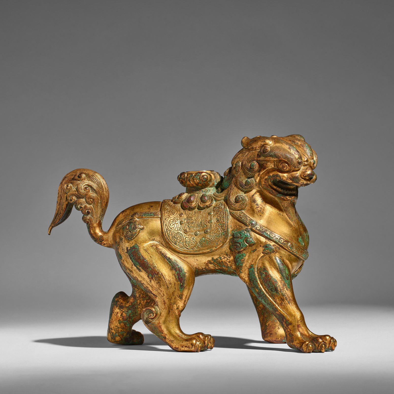 A RARE GILT-BRONZE BUDDHIST LION 10th century (blue brocade box): A RARE GILT-BRONZE BUDDHIST LION 10th century The ferocious beast striding with its head turning to the right, the bulging eyes glaring downward beneath the furrowed brow and tufts of curling fur, its