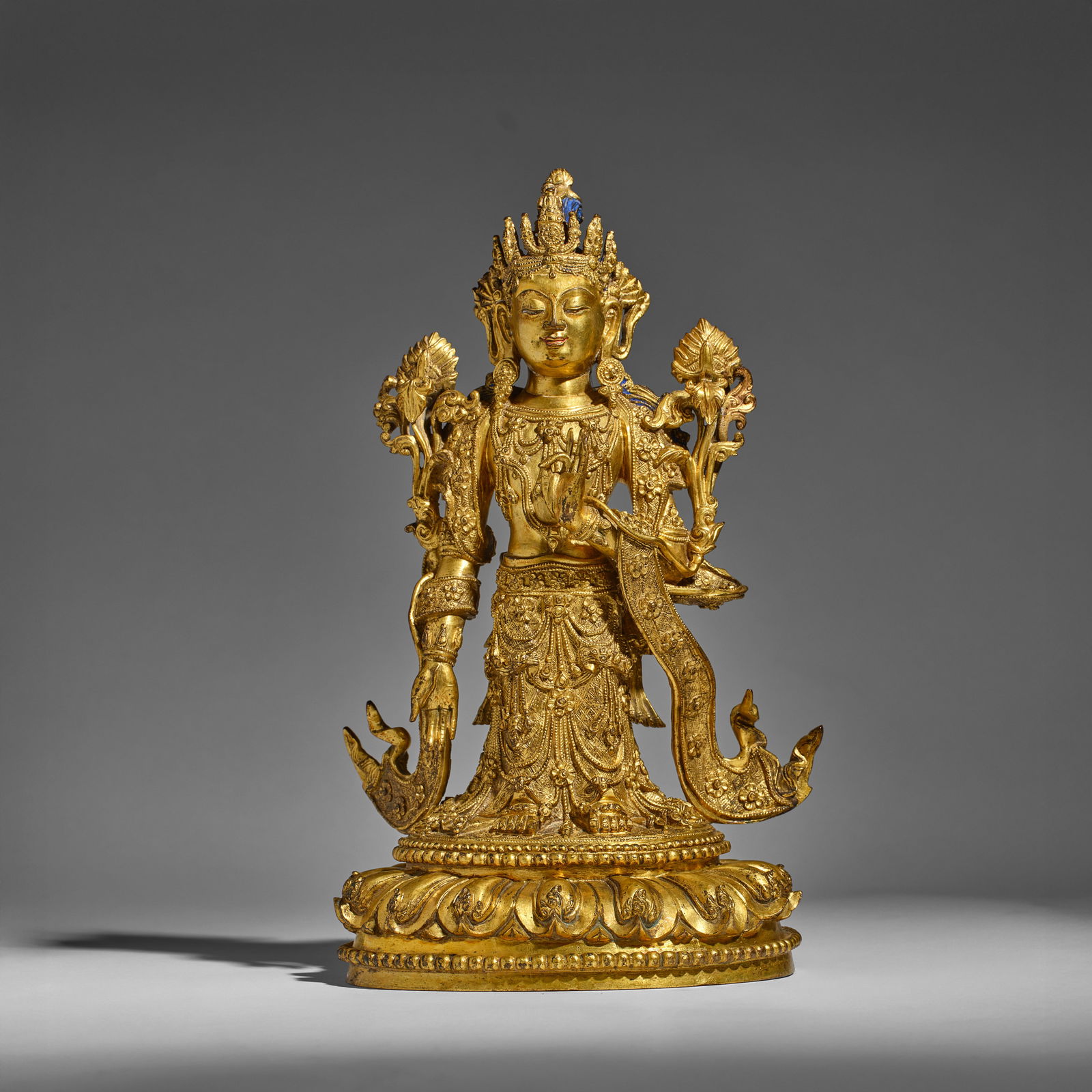 A GILT-BRONZE FIGURE OF BODHISATTVA AVAOLKITESVARA 16th/17th century (brocade fitted box): A GILT-BRONZE FIGURE OF BODHISATTVA AVAOLKITESVARA 16th/17th century Finely cast standing, the figure flanked at each shoulder by luxuriant blossoms and scrolling foliage, their stems curling into the