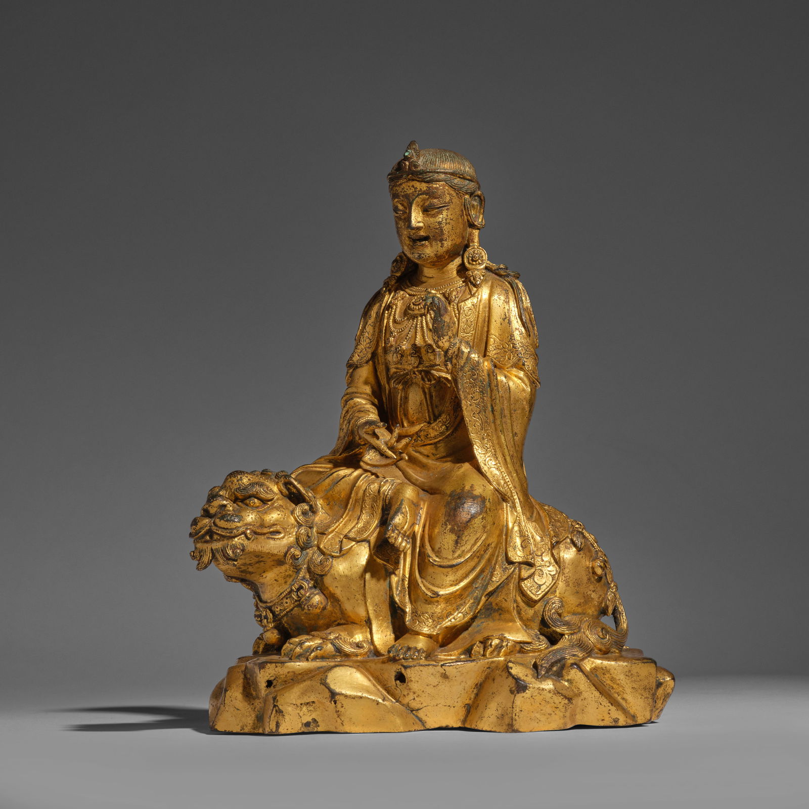 A GILT-BRONZE FIGURE OF MANJUSRI ON BUDDHIST LION Yuan/Early Ming Dynasty (brocade fitted box): A GILT-BRONZE FIGURE OF MANJUSRI ON BUDDHIST LION Yuan/Early Ming Dynasty Elegantly cast seated in lalitasana (the royal ease) atop a recumbent Buddhist lion, the left hand raised in vitarkamudra and