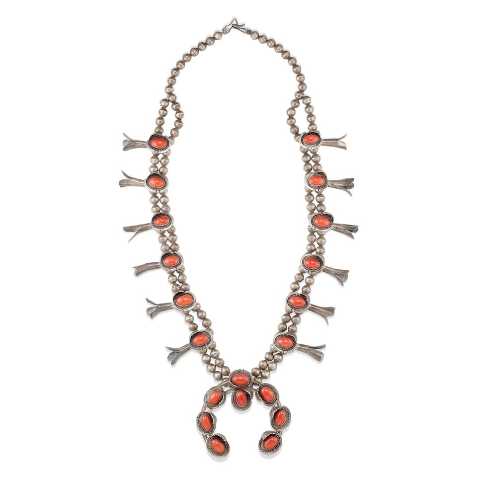 A Diné (Navajo) squash blossom necklace: A Diné (Navajo) squash blossom necklace Designed with two strands of silver beads strung with twelve coral-set blossoms, each with a feather-form silver accent, with a conforming naja set with ei