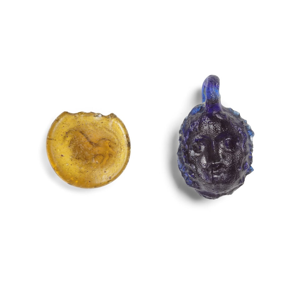 A Hellenistic dark blue glass janiform head pendant and a Roman amber glass token stamped with a... - 2