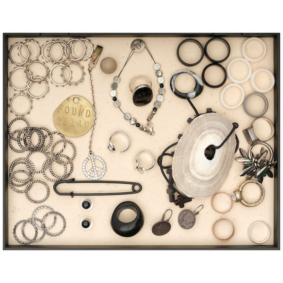 A CURATED BOX OF RUBBER, METAL, AND BEADED RINGS WITH FOUND OBJECTS: A CURATED BOX OF RUBBER, METAL, AND BEADED RINGS WITH FOUND OBJECTS Including an assortment two metal rings spelling DK, a black large safety pin, a wire beaded bracelet with the letter D, and a
