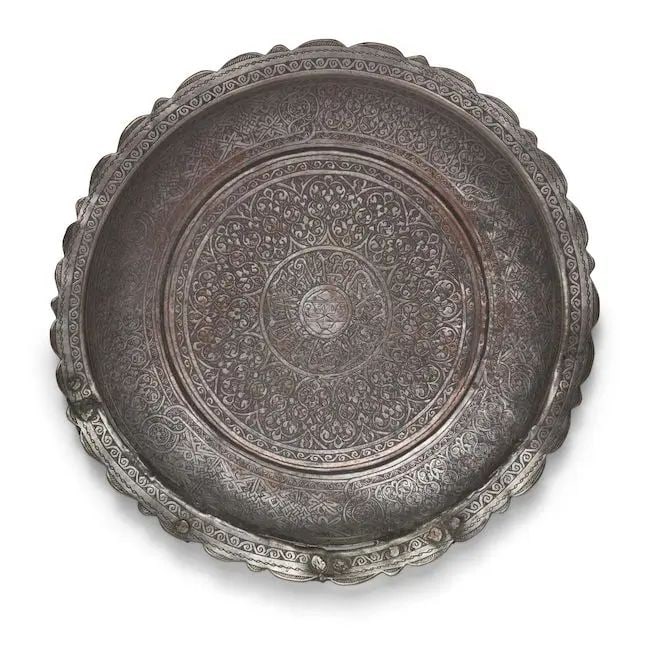 A Mamluk tinned-copper dish Egypt or Syria, early 16th Century: A Mamluk tinned-copper dish Egypt or Syria, early 16th Century of shallow circular form with lobed rim, profusely engraved with a central roundel filled with inscriptions in thuluth around a heraldic 