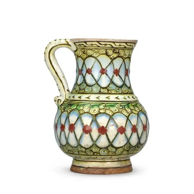 An Iznik pottery jug Turkey, circa 1600: An Iznik pottery jug Turkey, circa 1600 of piriform with waisted cylindrical neck and simple handle on a short splayed foot, decorated in raised-red, cobalt-blue, green and black outline on a white gr