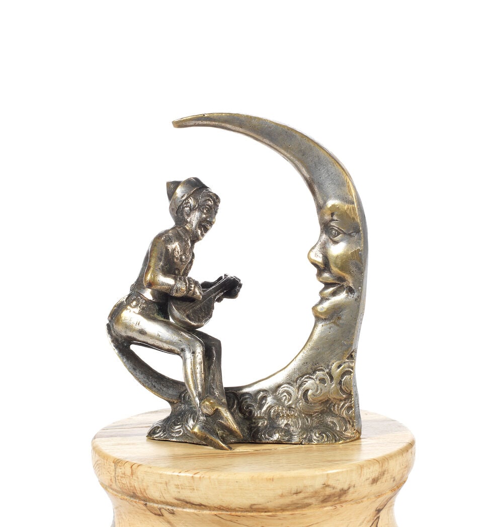 A 'Pierrot Serenading the Moon' mascot by Bioke, circa 1910,: A 'Pierrot Serenading the Moon' mascot by Bioke, circa 1910, nickel-plated brass, signed to left side of base, depicting a Pierrot with mandolin seated on a crescent moon, 10.5cm high, (some abrasion 