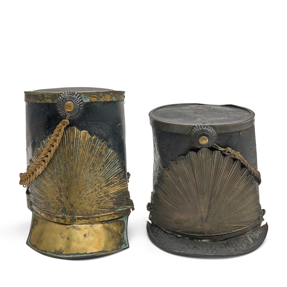 TWO 19TH CENTURY NEW ENGLAND MILITIA SHAKOS,: TWO 19TH CENTURY NEW ENGLAND MILITIA SHAKOS, c. 1810-1830. The black leather caps with embossed brass sunburst plates on the front above a brass or leather visor, eagle button mounting a gilt tassel, 