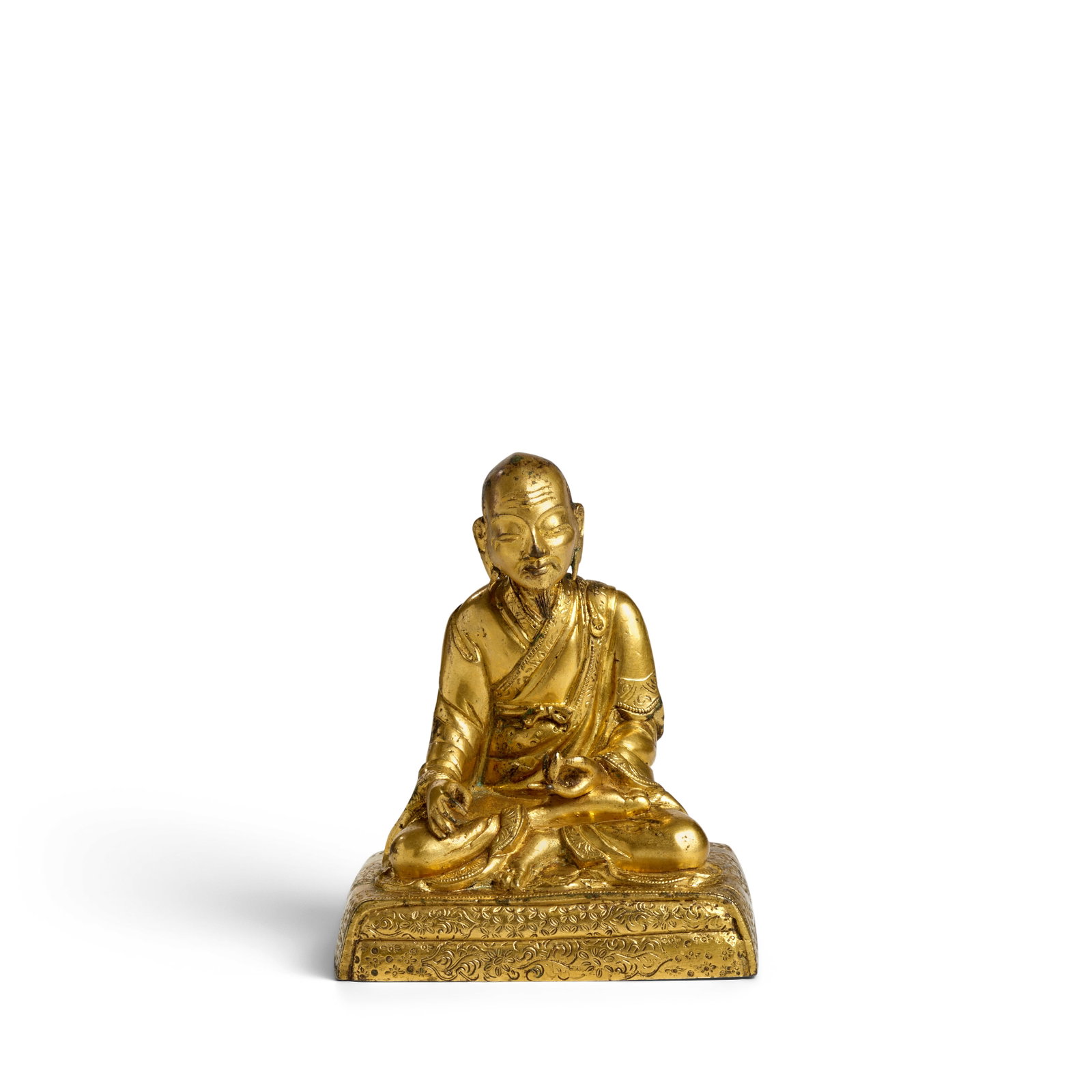 A SINO-TIBETAN GILT-BRONZE FIGURE OF A LAMA 18th century (fitted box): A SINO-TIBETAN GILT-BRONZE FIGURE OF A LAMA 18th century Seated in vajraparyanka on a rectangular carpeted throne finely engraved with floral scrolls and diaper borders, dressed in layered monastic ro