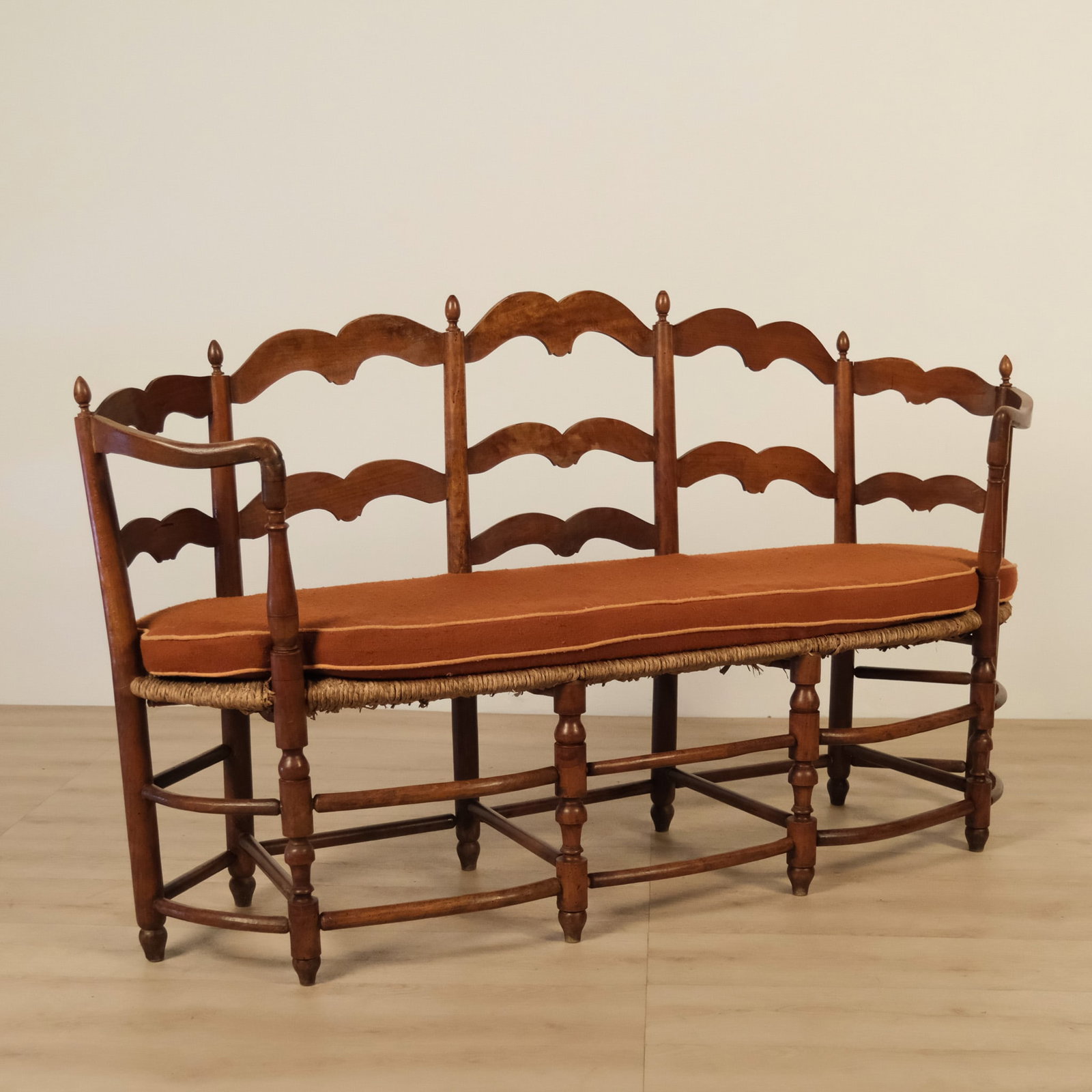 French Provincial Bench, c.1900: A charming antique French provincial settee, crafted in warm-toned fruitwood with a three-panel ladder back and woven rush seat. The tall, gently scalloped crest rail and matching shaped back slats