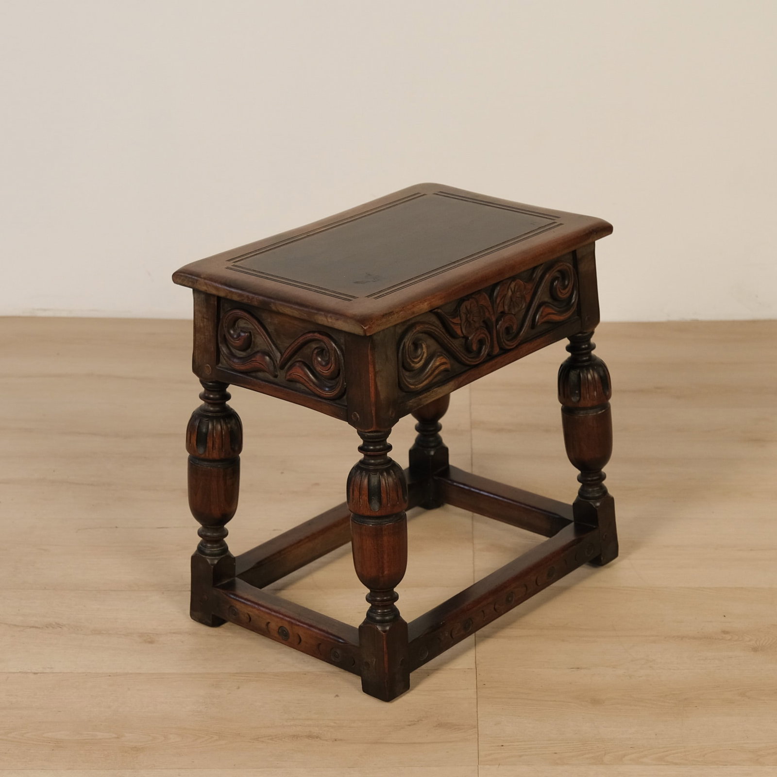 Carved Jacobean-style Oak Joint Stool: Jacobean-style joint stool featuring a rectangular top with a rich, dark patinated center panel framed by a tooled border, above a skirt carved in relief with scrolling foliate and floral motifs.