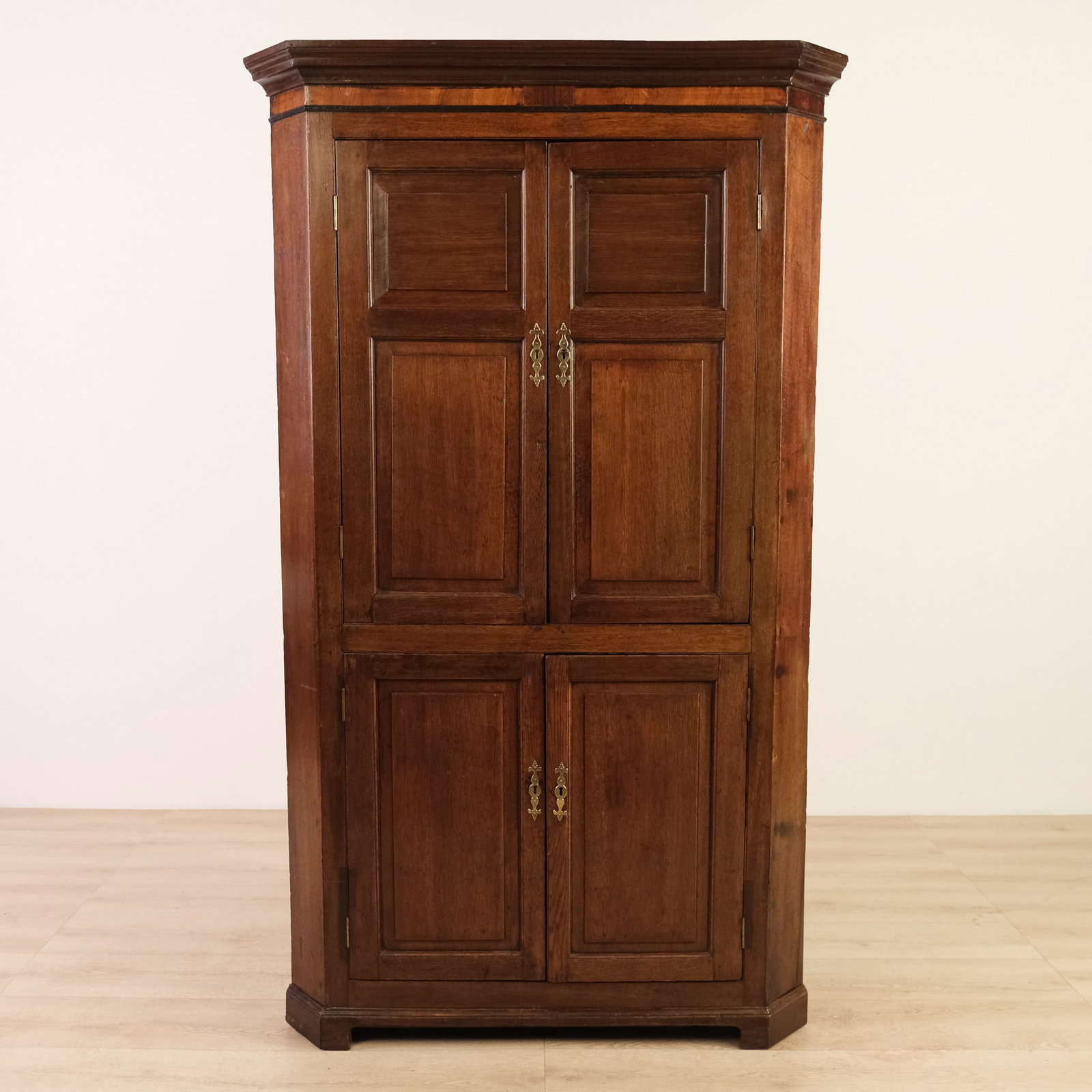 English or Continental Corner Cabinet, c. 19th Century: Tall 19th century standing corner cabinet with canted front corners, featuring a bold molded cornice above two sets of raised-panel doors each fitted with brass fleur-de-lis pull escutcheons. The