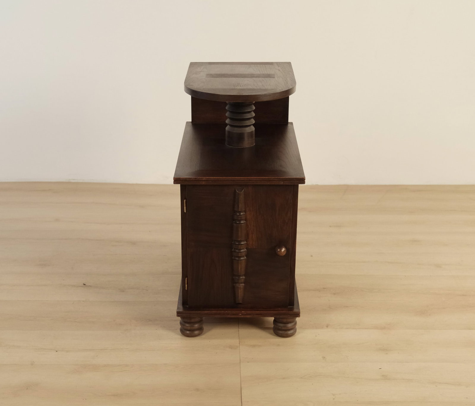 Charles Dudouyt Style Two-Tier Side Table with Cabinet Base: This Charles Dudouyt style end table features dark wood, bold geometric shapes, and distinctive carved accents. The piece has a sturdy cupboard base with a round knob and relief-carved column, a