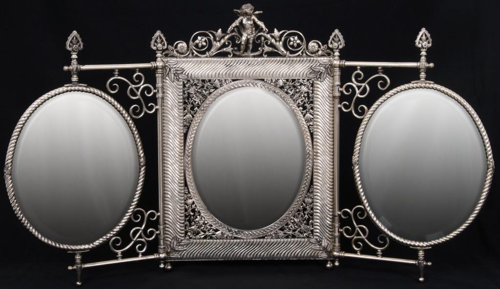 Exceptional Silver Plated Dressing Mirror: Three panel figural dressing mirror has an elaborate designed oval beveled glass center mirror with pierced decorated border having leaves, vines and flowers; above stands a winged cherub with two scr