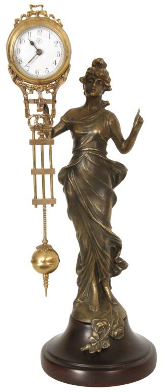 Junghans Figural Swinger Mystery Clock: Junghans mystery clock depicts a standing spelter figure of a woman wearing long garments. The gridiron pendulum rests on 2 pins on a pivot pointing forward from the figure's right hand; the pendulum 