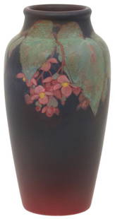 Rookwood Pottery Matte Vase: Rookwood Pottery vase is decorated with large green leaves encircling the top having branches with clusters of pink flowers hanging down; set against a deep purple background with red undercoat.