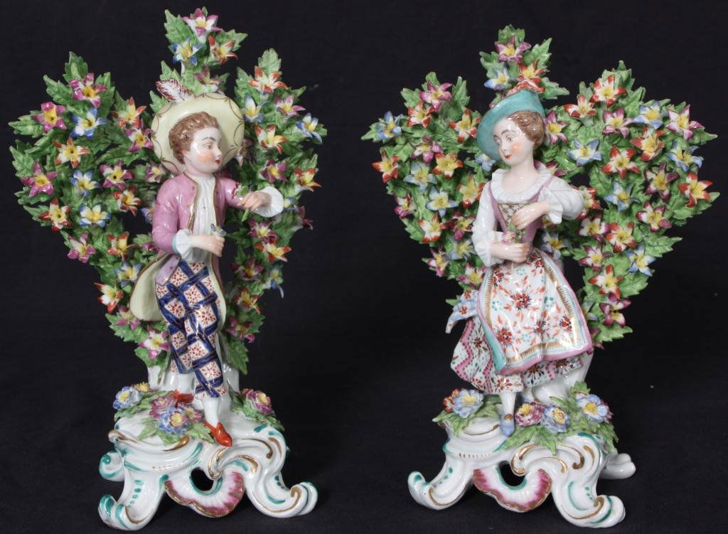 Pr. German Porcelain Figures: Pr. German Porcelain Figures. Figure of a man and figure of a woman both are wearing colorful traditional garments and hats and holding flowers in their hands; rising up behind them is a blooming wall