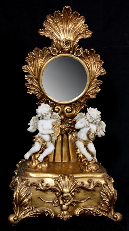 Gilt Carved Cherub Table Mirror: Gilt Carved Cherub Table Mirror. Figural pair of winged bisque musical cherubs, one playing a flute, one playing a lyre; a gilt carved framed mirror behind the figures has leaf and scroll design with 