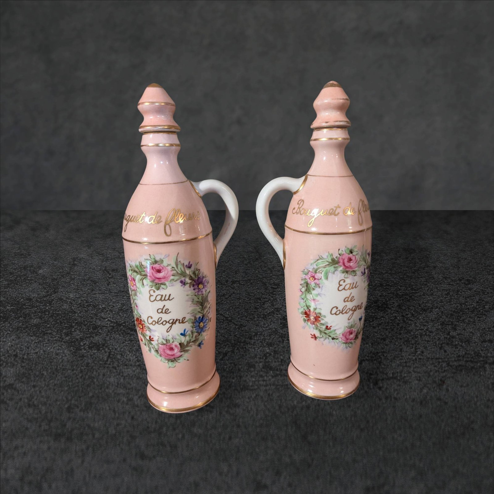 10 1/2 In. Lamoges Hand-Painted Porcelain Cologne Bottles: Porcelain cologne bottles measuring 10 1/2 inches tall, with pink hand-painted exteriors. Each features 'Eau de Cologne' text surrounded by floral garlands and gold-painted accents. Stoppered tops and