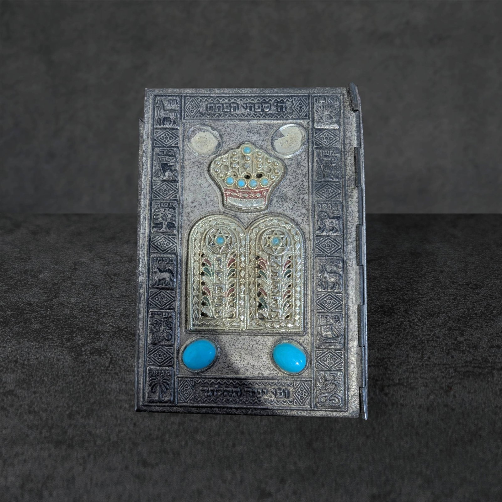 Silver-Plated Hebrew-English Siddur Prayer Book with Decorative Cover: A Hebrew-English siddur prayer book with a silver-plated cover featuring intricate designs, including a crown motif with turquoise-style accents on the front. The back cover displays embossed scenes, 