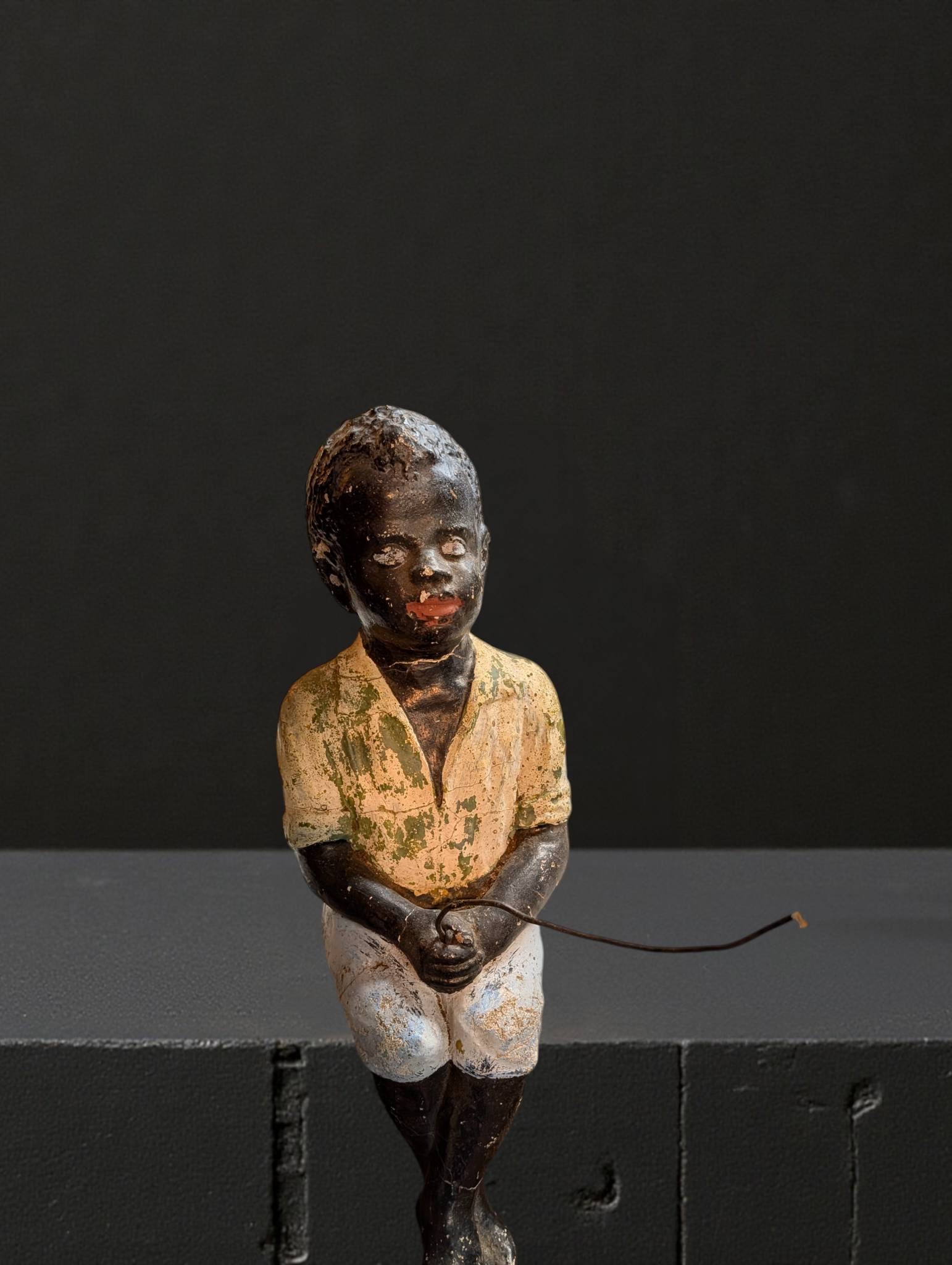 African American Concrete Figurine, 18 Inches Tall: Concrete sculpture depicting an African American figure seated, holding a thin rod or staff. Surface is painted with visible wear, including chipped paint on the yellow shirt, white pants, and skin-to