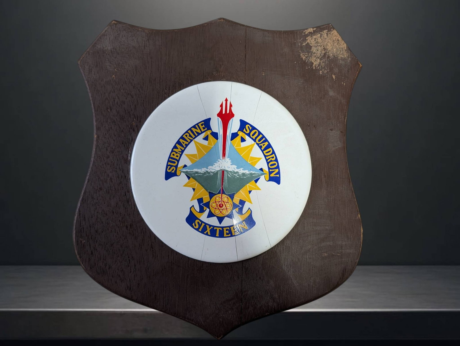 Submarine Squadron 16 Porcelain and Wood Plaque with Cracked Surface: Submarine Squadron 16 shield-shaped wooden plaque featuring a central porcelain inlay decorated with the squadron emblem, including a submarine and trident motif. The porcelain inlay has a visible cra