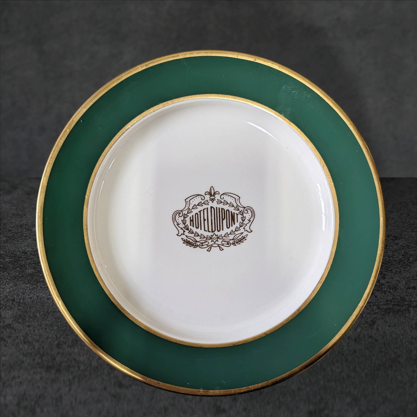 Hotel DuPont Plate by Mayer China, 9-Inch Diameter: A ceramic plate manufactured by Mayer China, featuring the iconic Hotel DuPont logo printed at the center of the dish. The plate has a green border accented with gold trim. The back of the plate bears