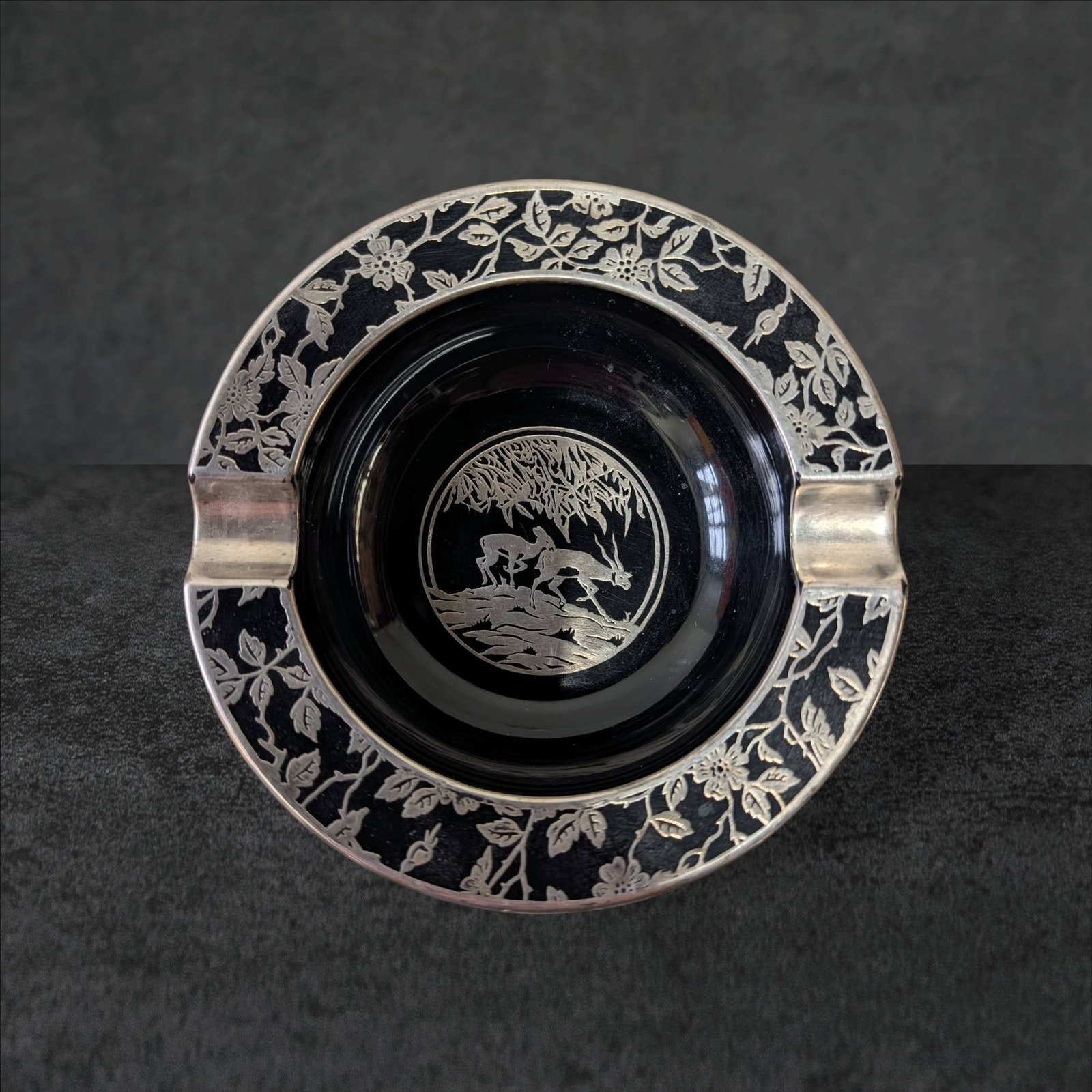 Black Amethyst Glass Ashtray with Silver Overlay Woodland Scene: Black amethyst glass ashtray featuring a detailed silver overlay depicting a woodland scene with deer. The circular dish measures approximately 5 inches in diameter, with decorative silver scrolling a