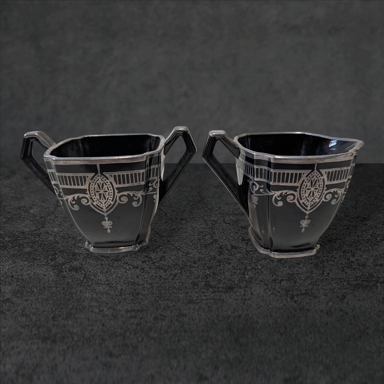 Black Amethyst Sugar Bowl and Creamer with Silver Overlay Deco Motif: A black amethyst glass sugar bowl and creamer set featuring a silver overlay with an Art Deco motif. The pieces measure approximately 3.5 inches tall and 4.5 inches wide. Each piece has geometric hand