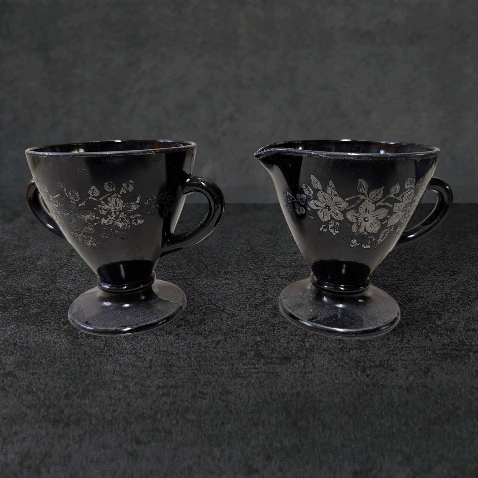 Amethyst Glass Cream and Sugar Set with Silver Overlay: Cream and sugar set crafted from amethyst-colored glass, featuring decorative silver overlay floral motifs. Both pieces are approximately 3 1/2 inches tall and 4 inches in diameter. The cream pitcher 
