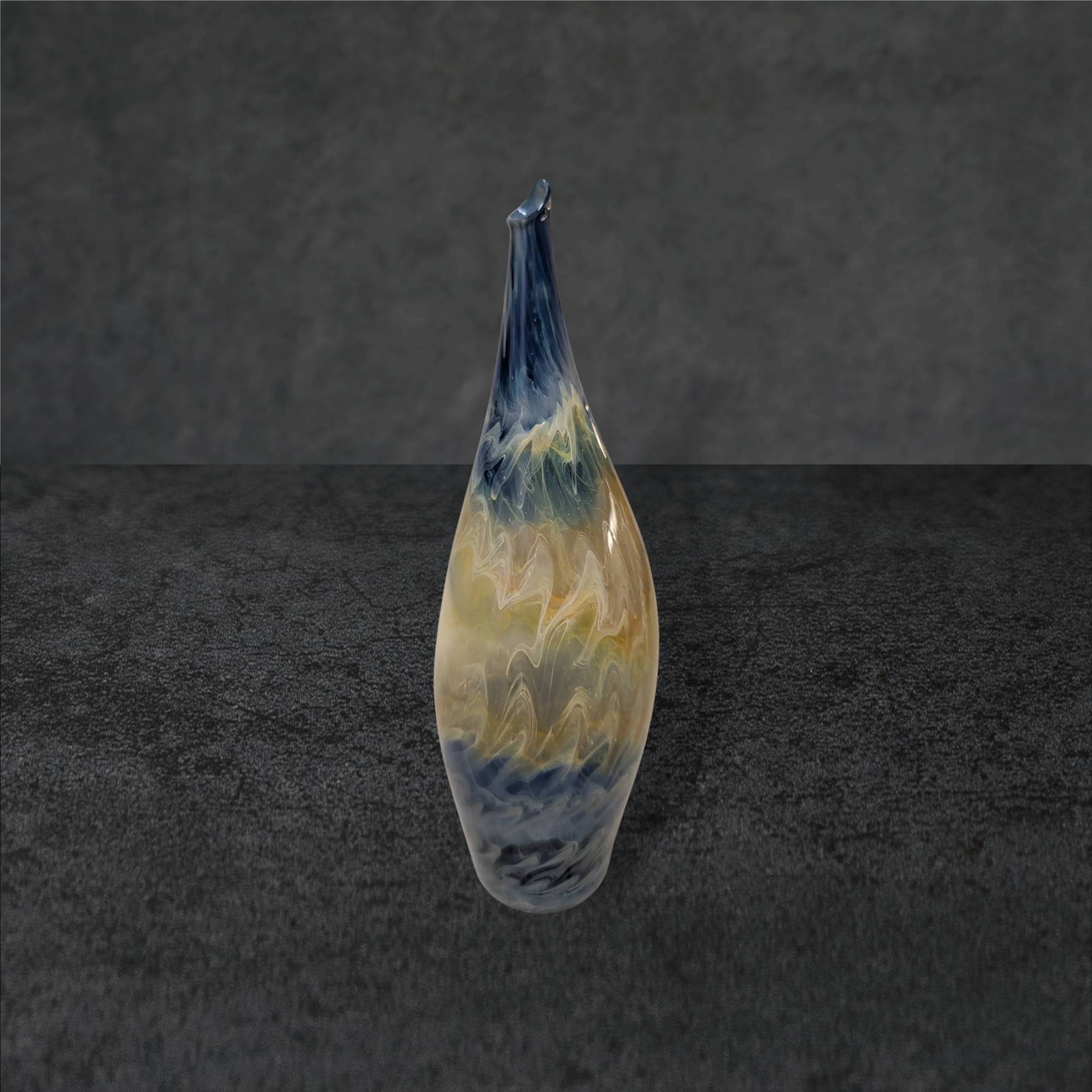 Hem Blown Glass Vase, 7 1/2 Inches Tall: Blown glass vase attributed to Hem, standing 7 1/2 inches tall. Features a tapered form with a slightly curved, pointed tip. The vase displays a swirling multicolor design, predominantly blue, yellow,