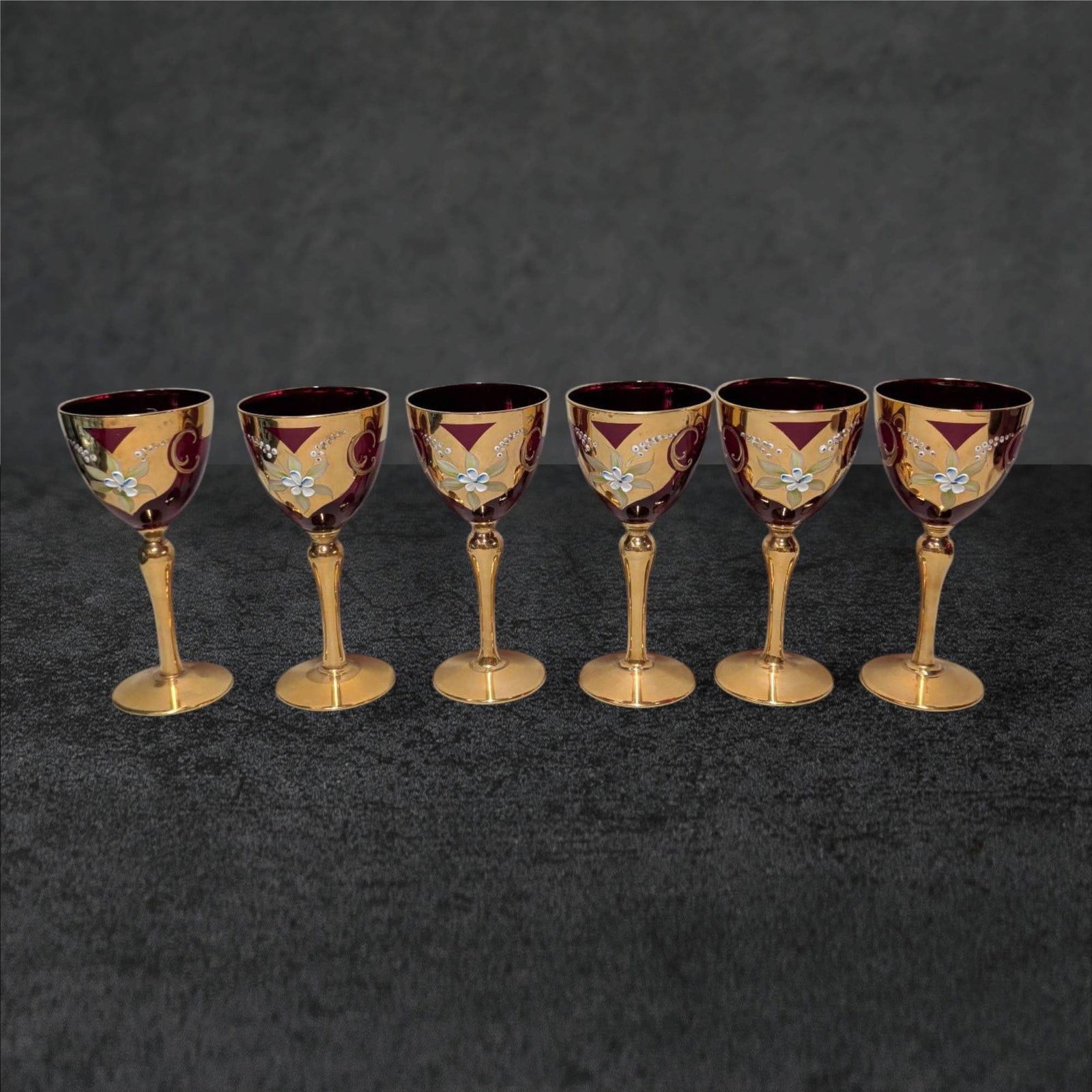 Six Murano Red and 24K Gold Wine Glasses with Hand-Painted Floral Design: Six handcrafted Murano wine glasses featuring deep red bowls adorned with 24K gold accents and floral hand-painted decorations. Each glass has a gold-toned stem and base, embodying mid-20th-century Ve