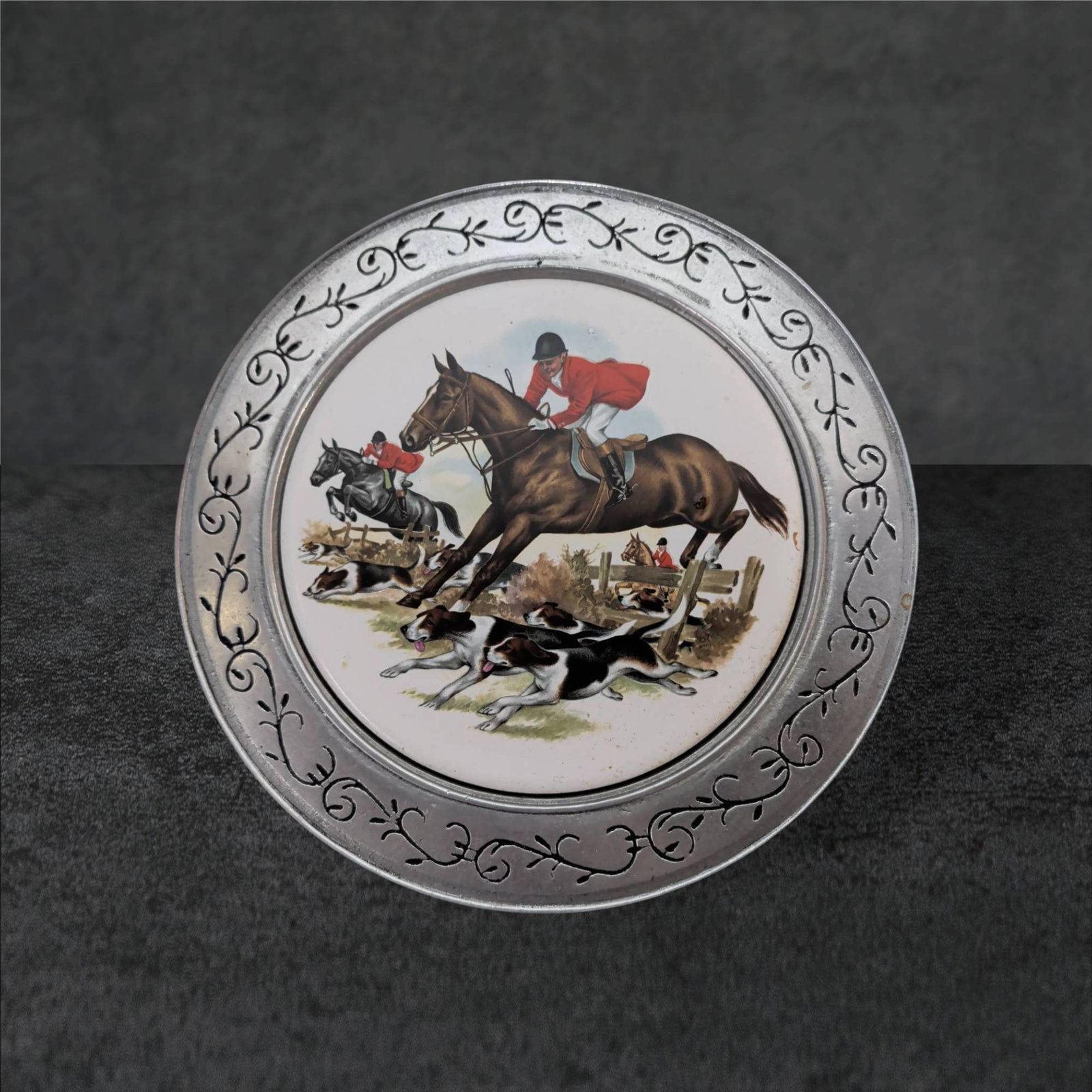 Round Metal Trivet with Porcelain Hunt Scene, 8.5-Inch Diameter: Circular trivet featuring a porcelain center depicting a hunt scene with riders on horseback and dogs. The metal border includes an engraved scrollwork design. Back side is flat and includes a small h