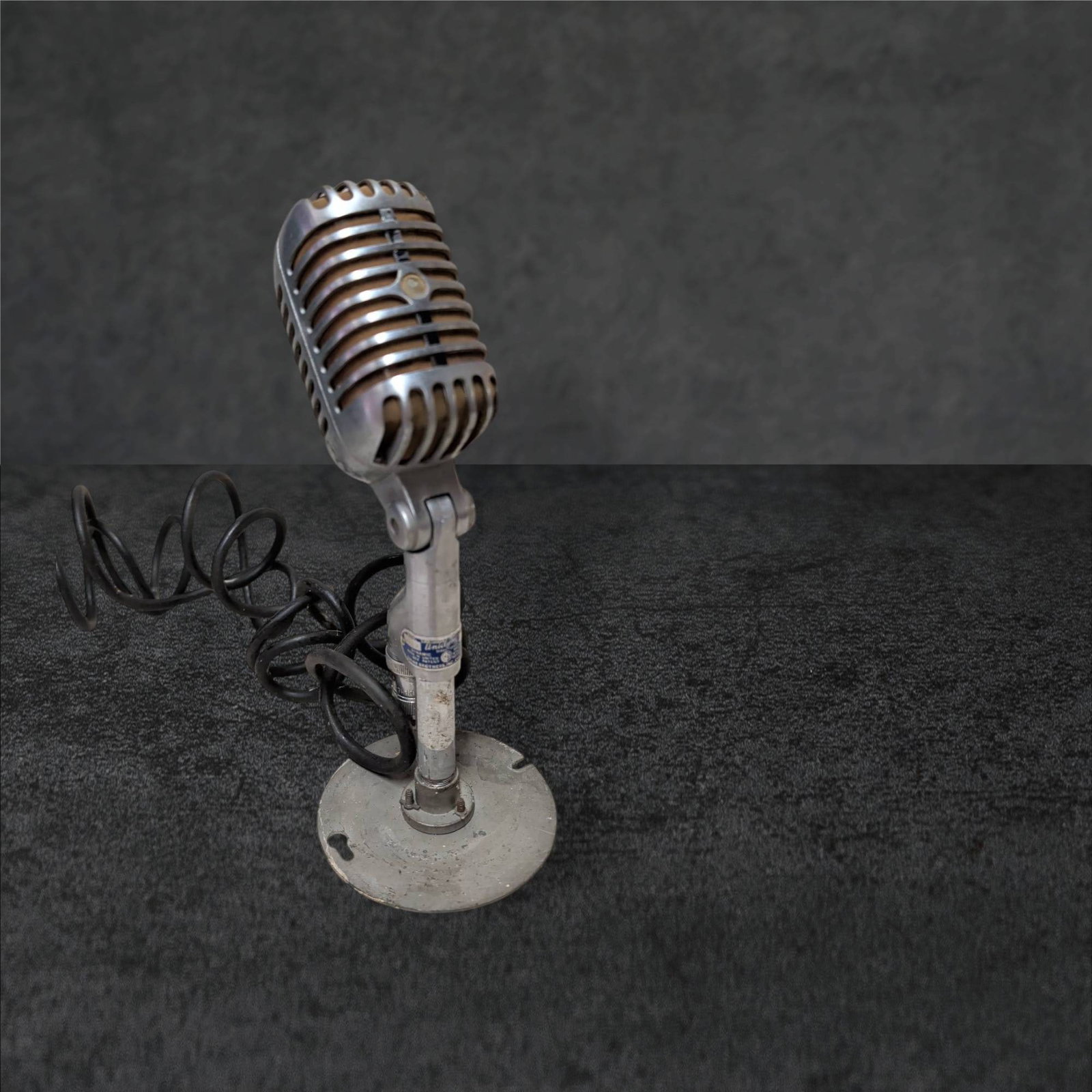 Univine Supercardioid Dynamic Microphone Without Badge, 10 in. Tall: Univine supercardioid dynamic microphone measuring approximately 10 inches in height. Constructed with a metal grille and integrated stand. The microphone shows wear and patina, with a missing front b