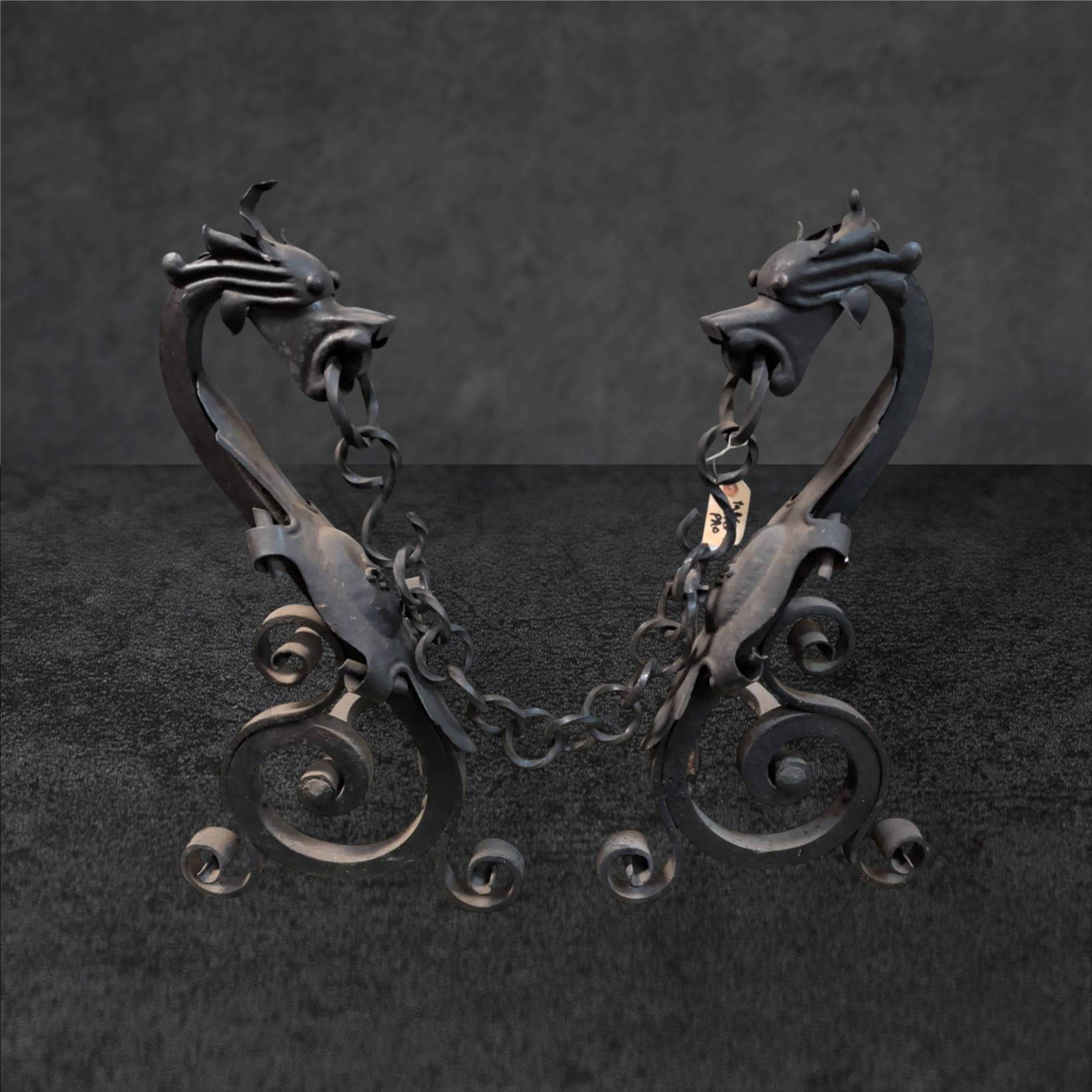 Wrought Iron Dragon Andirons, 28 Inches Tall: Wrought iron andirons featuring dragon motifs, each measuring 28 inches tall, 14 inches wide, and 28 inches long. The andirons display elaborate scrollwork and chains held in the dragons' mouths. Cons
