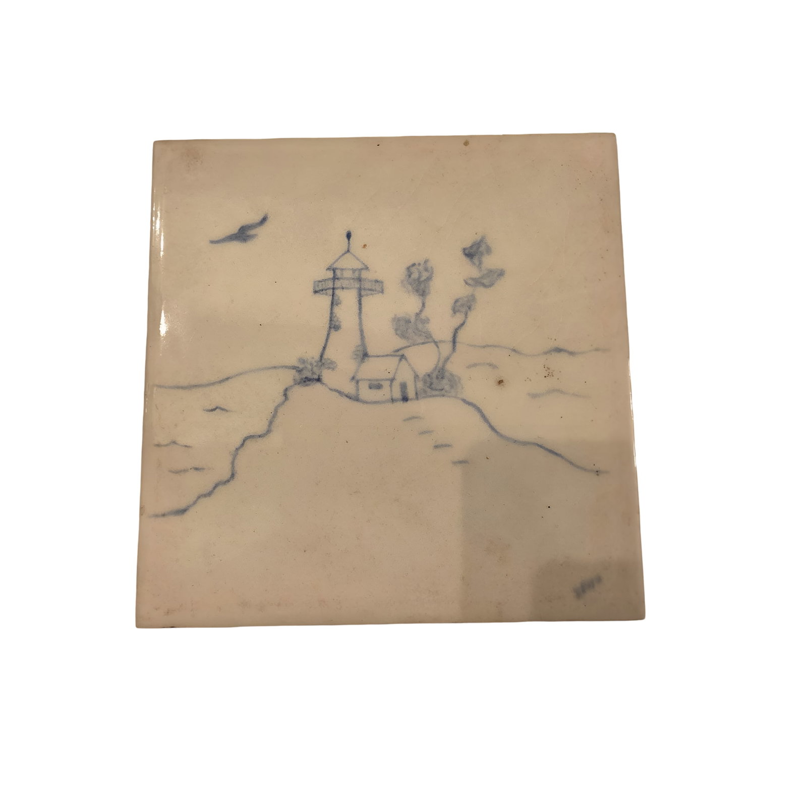 Decorative Ceramic Tile with Lighthouse and Coastal Scene: Ceramic tile featuring a hand-painted blue coastal scene, including a lighthouse, small building, trees, and a bird in flight. The edges are straight, and the back shows signs of wear consistent with