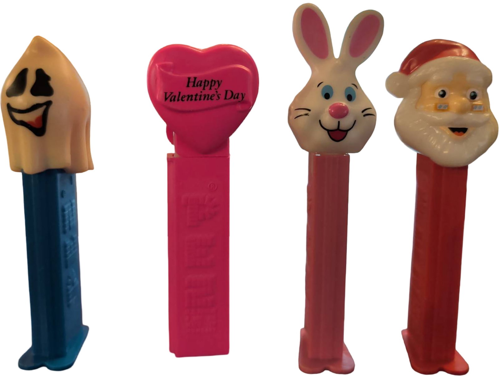 Four Pez Dispensers Featuring Various Themed Characters: Four Pez candy dispensers with character tops: ghost with blue base, pink heart inscribed 'Happy Valentine's Day,' white bunny with pink base, and Santa Claus with red base. All dispensers feature sta